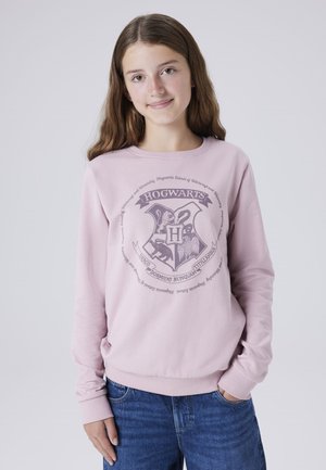 Roza pulover s kratkimi rokavi, ki ima v sivi barvi grafiko grba Hogwartsa. Mehki bombažni material z rebrastimi manšetami in robom. Nosi se z modrimi kavbojskimi hlačami.