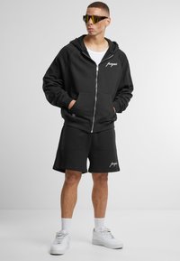 Musta vetoketjullinen huppari, jossa on logo, yhdistettynä mustiin shortseihin. Molemmat ovat puuvillaa ja niissä on rento istuvuus. Valkoiset tennarit täydentävät asustusta.