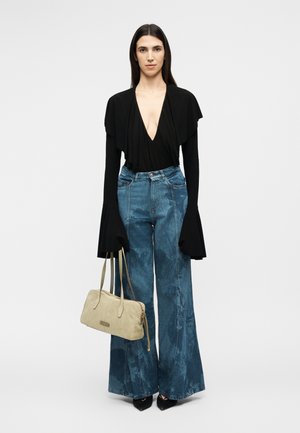 Ženska stoji in nosi črno dolgo majico z globokim V-izrezom ter razširjenimi manšetami, modre kavbojke z visokim pasom in širokimi hlačnicami, črne čevlje in se drži bež torbice.