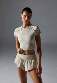 Mladá žena s tmavými vlasmi v hladkom copu, oblečená v béžovom priliehavom crop tope a zodpovedajúcich šortkách s vysokým pásom, na neutrálne šedom pozadí.
