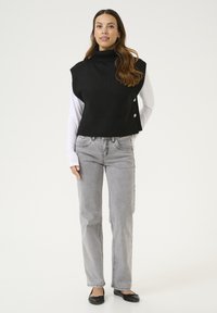 Čierny pletený bezrukávový rolák preložený cez bielu košeľu s dlhým rukávom, v kombinácii so svetlošedými rovnými džínsami a čiernymi plochými topánkami.