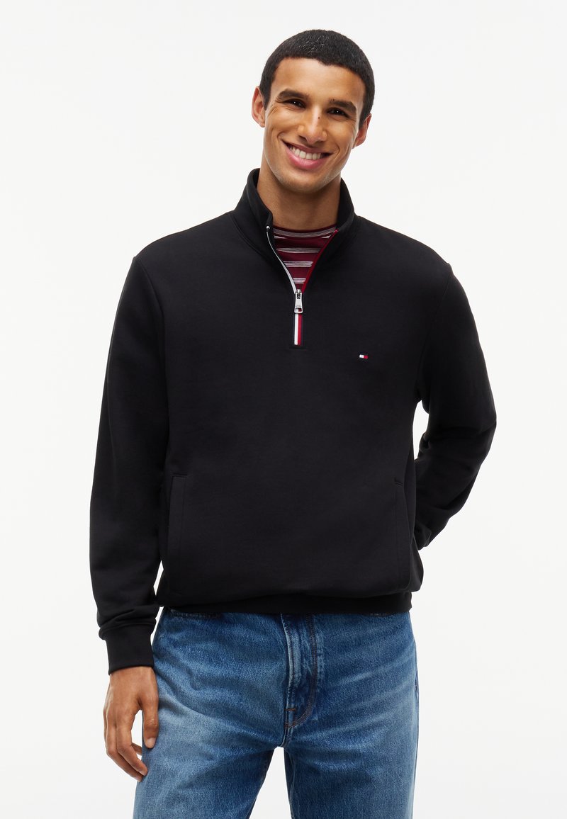 Homme souriant portant un pull noir à demi-fermeture éclair sur une chemise rouge rayée et un jean bleu, debout devant un fond blanc.