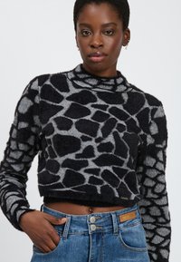Giovane donna con un pullover cropped a fantasia giraffa nero e grigio e jeans a vita alta blu, con una mano in tasca, su uno sfondo neutro.