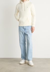 Sudadera con capucha de color crema claro con bolsillo frontal, combinada con jeans rectos de color azul claro y zapatillas blancas sobre un fondo neutro.