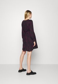 Vestido preto com padr�ão de folhas roxas, mangas longas, cinto amarrado na cintura, comprimento até ao joelho. Usado com sapatos pretos de slip-on. Fundo simples.