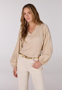 Beige langemouwtop met een V-hals en textuurstof, gecombineerd met lichte broek en een gouden riem. Zachte, relaxte pasvorm met pofmouwen.