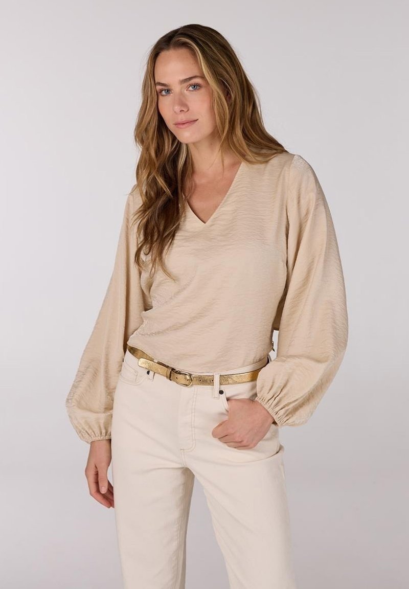 Beige langemouwtop met een V-hals en textuurstof, gecombineerd met lichte broek en een gouden riem. Zachte, relaxte pasvorm met pofmouwen.