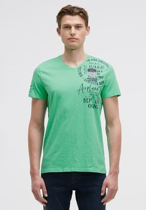 Young man wearing a green V-neck T-shirt with black printed text and graphics on chest and left sleeve, paired with dark blue jeans.