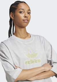 Pelēks lielgabarīta t-krekls ar īsām piedurknēm, kas piedāvā teksturētu dzeltenu un baltu izšūtu Adidas logo uz krūtīm.