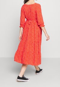 Vestido rojo con lunares, de corte relajado, mangas largas y falda de volantes, ajustado en la cintura con una cinta de tela. Combinado con zapatillas negras.