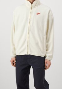 Krämfärgad fleece-jacka med dragkedja och hög krage, samt röd Nike-logotyp på vänster bröst. Har sidofickor och ribbade manschetter.