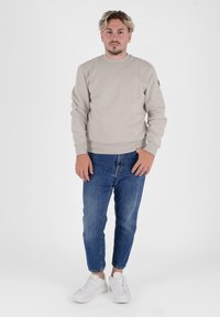 Beżowa bawełniana bluza z okrągłym dekoltem, ściągaczami przy mankietach i u dołu. Połączona z niebieskimi, dopasowanymi dżinsami i białymi sneakersami. Prosty design, bez wzorów.