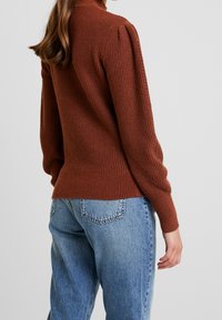 Roodbruine geribde trui met een hoge kraag en pofmouwen, gecombineerd met lichtblauwe, hooggesneden jeans. Zijaanzicht weergegeven.