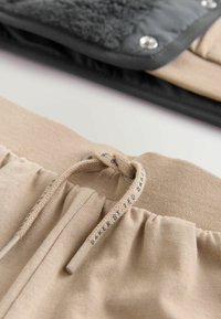Beige elastický pas s tkaničkou, vyrobený z texturované tkaniny a s vyztuženou průvlečkou. Pozadí zahrnuje černý a šedý oděv.