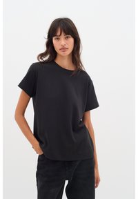 Mustast puuvillast lühikeste varrukatega ja ümara kaelusega T-särk. Lihtne, mugav lõige. Kandatud tumedate denimpükstega, hele taust.