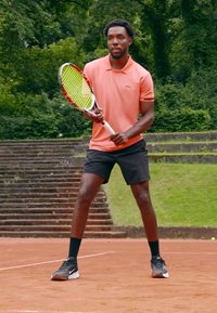 Tennisspiller som holder et grønt og rødt racket, iført en rosa polo-skjorte og svarte shorts, stående på en leirbane med grønn bakgrunn.