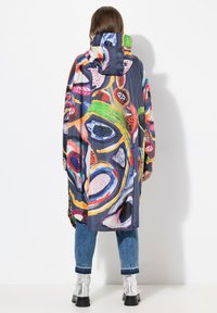 Poncho da pioggia colorato e fantasia, realizzato in materiale leggero con cappuccio. Presenta forme astratte in rosso, blu, verde e giallo su uno sfondo scuro.