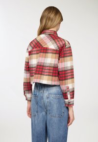 Giacca corta a quadri rossi e beige con maniche lunghe, dettaglio con bottoni sui polsini e collo strutturato. Indossata con pantaloni di jeans a vita alta.