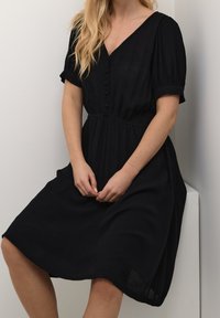 Schwarzes Kleid aus strukturiertem Stoff, mit V-Ausschnitt, kurzen Ärmeln, Knopfleiste und gerafftem Taillenbereich, sowie einem schwingenden A-Linienrock.