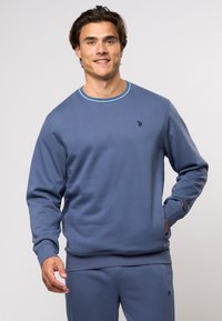 Modra dukserica z dolgimi rokavi, rebrastim okroglim izrezom in kontrastnimi šivi. Ima majhen logotip na prsih in gladko teksturo.