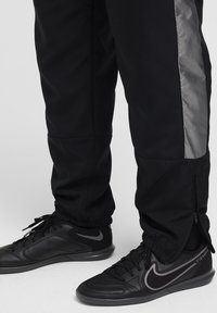 Schwarze Sporthose mit silbernem Seitenpanel und Reißverschluss an den Bündchen, kombiniert mit schwarzen Sneakers mit grauem Logo und strukturierten Akzenten.