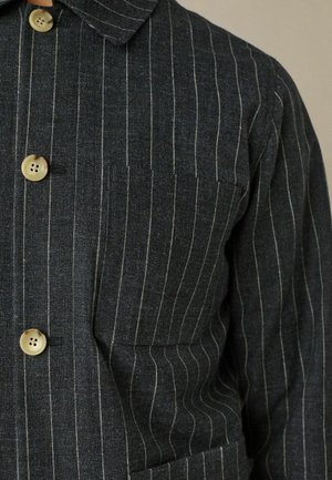 Camisa de botón gris oscuro con rayas verticales blancas, cuello con solapa y tres botones visibles en la parte delantera. Cuenta con un bolsillo en el pecho.