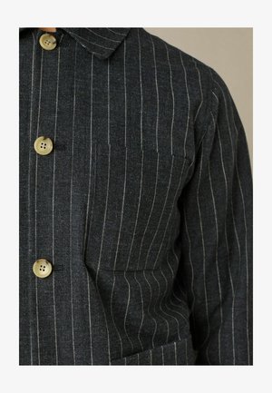 Temno siva srajca s kratkimi rokavi in navpičnimi belimi črtami, s ovratnikom in tremi vidnimi gumbi na sprednji strani. Vsebuje žep na prsih.