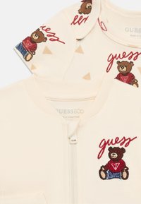 Krém színű gyermekruha cipzáras lezárással, hímzett medve motívummal és "guess" felirattal. A mintázat háromszögeket tartalmaz.