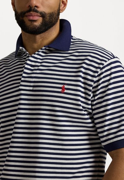 Marineblå og hvit stripete polo skjorte med en ensfarget marineblå krage. Har en liten rød brodert logo på brystet. Laget av strukturert stoff.