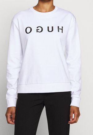 Person trägt ein weißes Sweatshirt mit langen Ärmeln und schwarzem, umgekehrtem "HUGO"-Schriftzug auf der Brust sowie schwarze Hose, an den Schultern beschnitten.