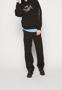 Czarna bluza z kapturem z nadrukiem i troczkami, nałożona na jasnoniebieską koszulę. Czarne spodnie typu cargo z bocznymi kieszeniami oraz czarne buty.