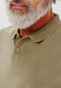 Khaki polo z żebrowanym kołnierzykiem, z dwoma guzikami i gładką fakturą. Noszony przez osobę z szarym zarostem, odsłaniającą górną część klatki piersiowej i szyję.