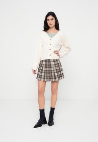 Κρεμ ζακέτα με χρυσά κουμπιά, σετ με καφέ καρό φαρδιά φούστα. Το μοντέλο φοράει ναυτικά καλτσάκια και μαύρα παπούτσια, στέκεται μπροστά σε απλό φόντο.