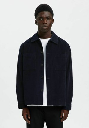 Joven vestido con una chaqueta de botonadura en sarga de color azul marino oscuro sobre una camisa blanca y pantalones negros, de pie contra un fondo claro y liso.