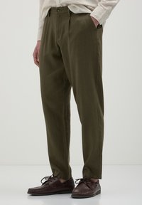 Pantalons en lin vert olive, à coupe ample, avec une texture lisse, des plis sur le devant et une fermeture à bouton ; assortis à des chaussures à lacets marron.