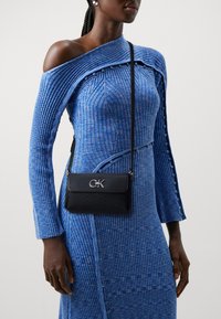 Borsa a tracolla in pelle nera con hardware argentato e logo CK, indossata con un vestito blu a spalla scoperta con righe verticali.