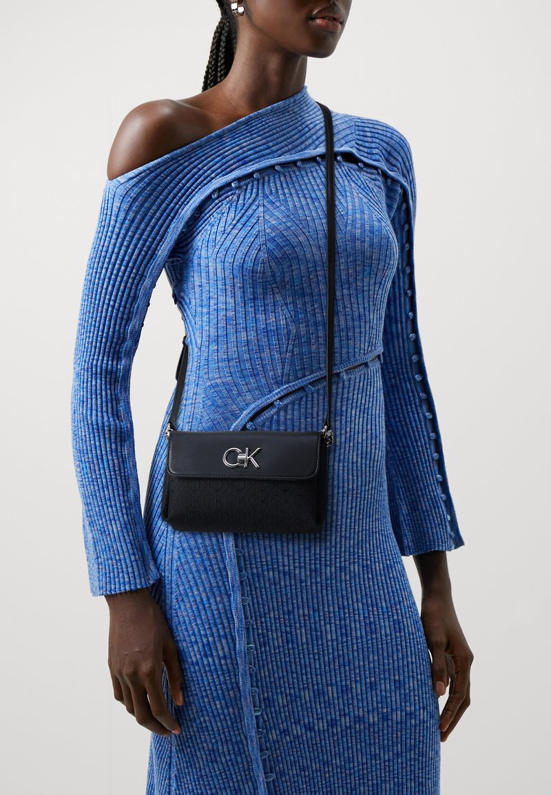 Borsa a tracolla in pelle nera con hardware argentato e logo CK, indossata con un vestito blu a spalla scoperta con righe verticali.