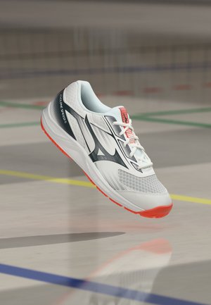 Fehér és fekete sportcipő narancssárga talppal és "Cyclone Speed 5" szöveggel, egy beltéri pálya padlója felett lebeg, amely színes vonalakkal van jelölve.