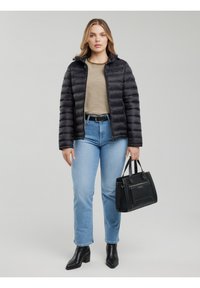 Sieviete, valkājot melnu pufīgu virsjaku virs svītrainas krekla un gaiši zilu džinsu biksēm, turot melnu somu, stāv uz vienkārša pelēka fona.