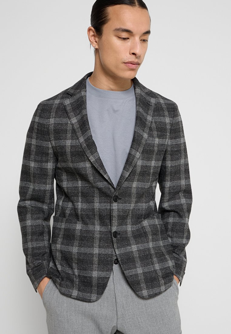 Mann in grauem karierter Blazer über einem hellgrauen Rundhalshemd mit den Händen in den Taschen, nach unten schauend vor einem schlichten Hintergrund.