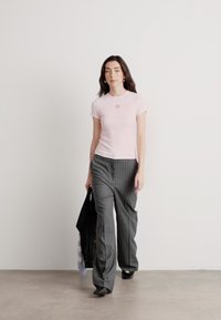 Heled roosa sobiv T-särk, millel on rinnal väike logo, koos hallide peenriidest laia säärega pükstega. Mudel hoiab musta kotti.