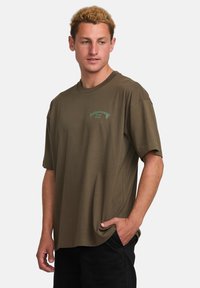 Olive green cotton t-shirt with short sleeves, round neckline, and a small logo graphic in light green on the left chest area.