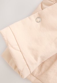 Veste matelassée beige clair avec tissu lisse, dotée d'un détail à bouton-pression argenté et de coutures matelassées, présentant une finition douce et texturée.