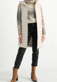 Osoba nosząca beżową, długą kamizelkę puchową, sweter z golfem w odcieniach jasnobrązowym i szarym, czarne dżinsy oraz beżowe botki na obcasie.