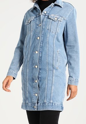Lichtblauw lange spijkerjas met knoopsluiting aan de voorkant, borstflappenzakken, zijzakken, gedragen over een zwarte top en broek.