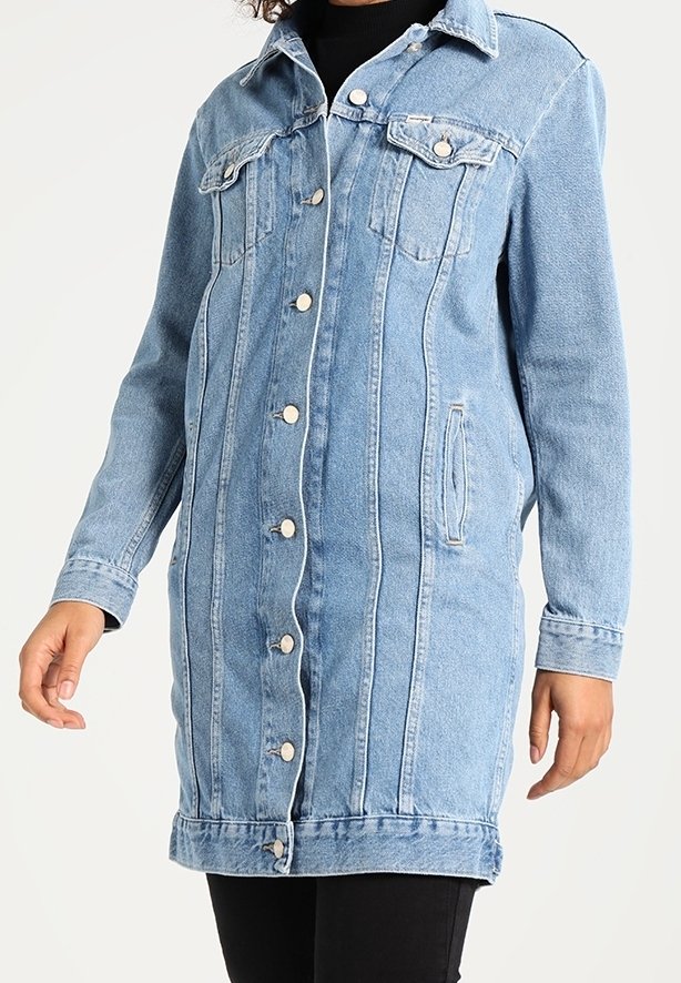 Veste longue en jean bleu clair avec fermeture à boutons sur l'avant, poches poitrine à rabat, poches latérales, portée sur un haut et un pantalon noirs.