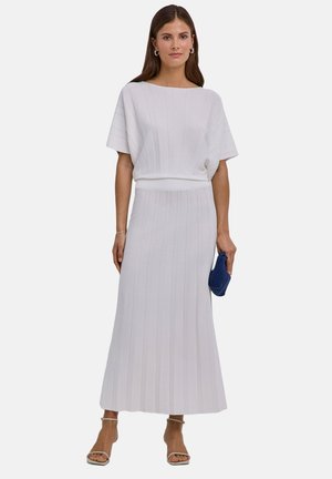 Vrouw die een wit geribd gebreid topje en bijpassende lange rok draagt, een kleine blauwe geweven clutch vasthoudt en witte sandalen met enkelbandje draagt.