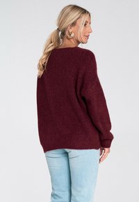 Kobieta z blond włosami w kucyku, nosząca duże złote kolczyki, bordowy sweter z dzianiny i jasnoniebieskie dżinsy, stojąca na tle jednolitego tła.