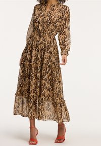Femme portant une robe à manches longues, à volants, marron et noire avec imprimé animal, associée à des sandales rouges à talons et à lanières.