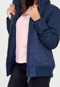 Mörkblå vadderad jacka med huva, med vertikala ränder, ribbade muddar och en dragkedjeficka, bärs över en ljusrosa topp.